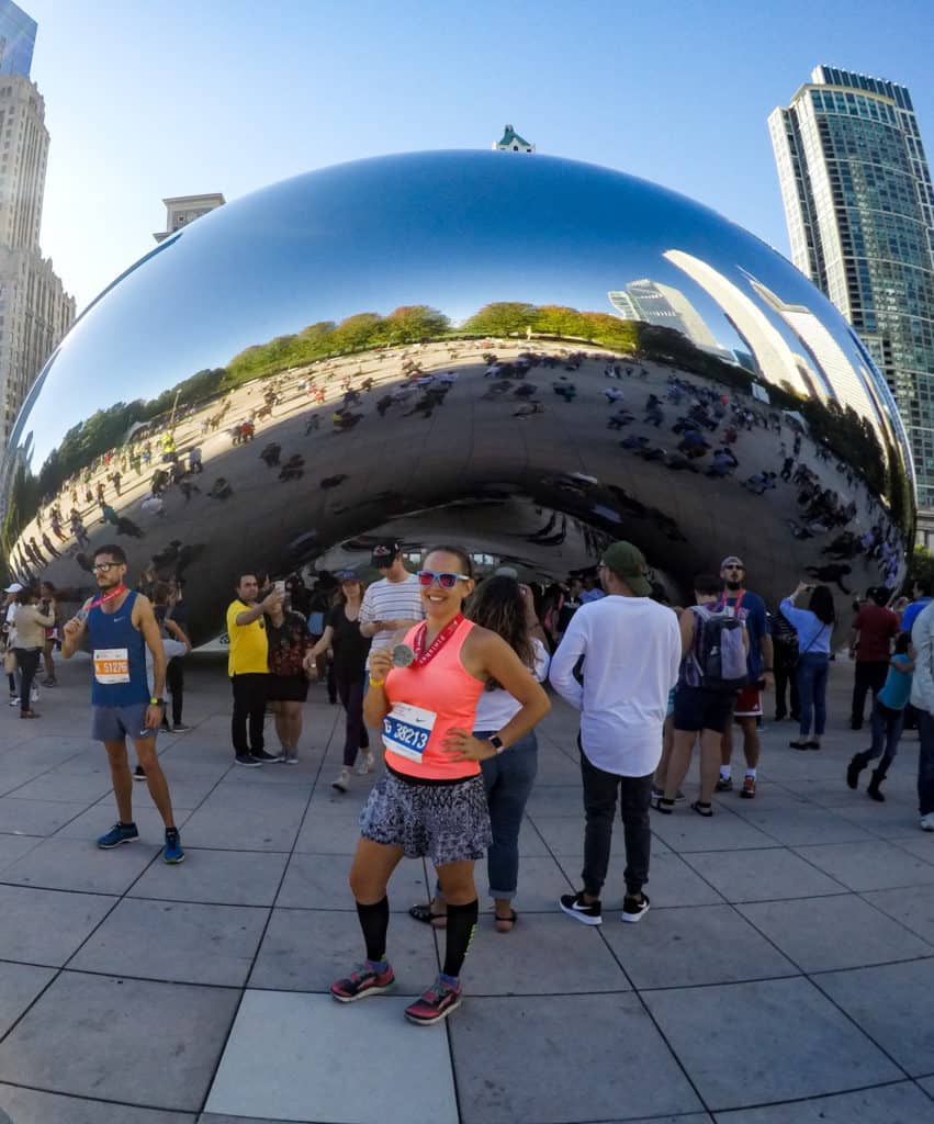 Female marathon runner poses in front of the Cloud Gate art installation in Chicago while holding a Chicago Marathon Medal. Picture is post-race of learning how to run walk a marathon.