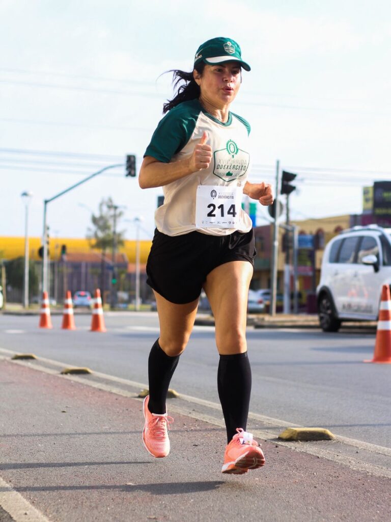 A woman is running a road race displaying a runner's bib with the number 214. Runners may ask, "how many steps are in a 5k?" Though the answer changes based on the runner, the estimate is around 5,000 steps.