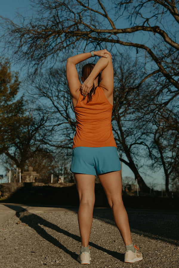 A female runner stretches the tricep muscle of her right arm with her back to the camera.