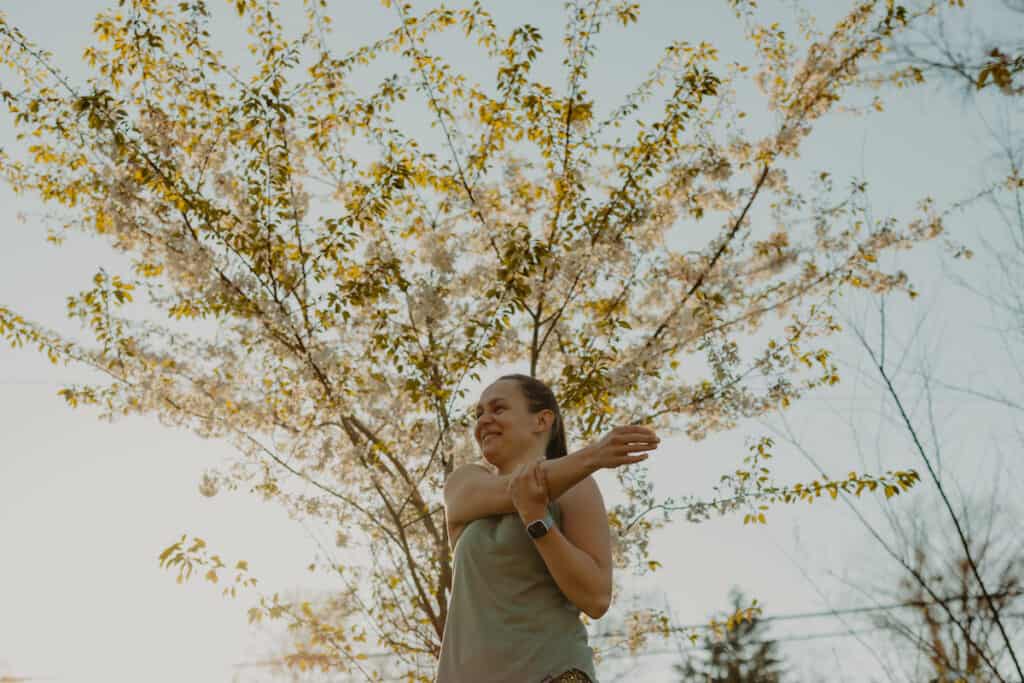 I am stretching my right arm by pulling it across my chest and to the left with my left hand. I am smiling as I stretch in front of a tree. Stretching is a great running tip for beginners.