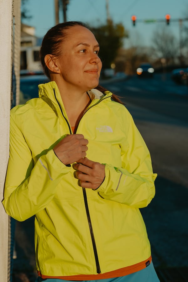 I'm smiling while leaning up against a build in my running rain jacket. I'm looking off to my left into the distance.