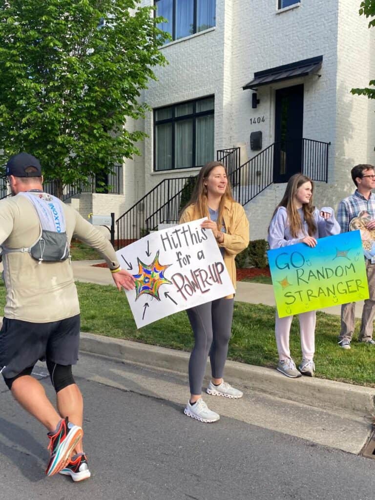 A marathon runner is passing by a spectator holding a sign that reads, "Hit this for a power-up." The runner is tapping the power-up with his hand. A spectator beside the first spectator holds a sign reading, "Go random stranger."