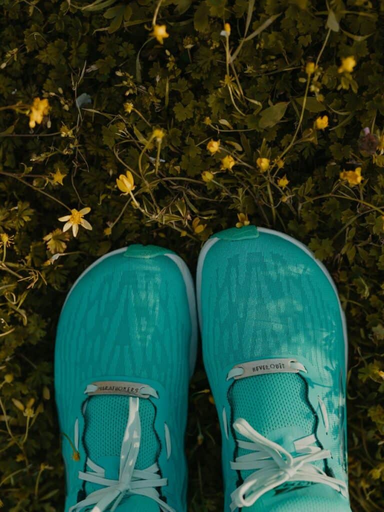 My Altra running shoes are in the grass. There are some small yellow flowers surrounding them. Looking from first person view there are lace charms that read "marathoners" on the left foot and "Never quit" on the right foot.