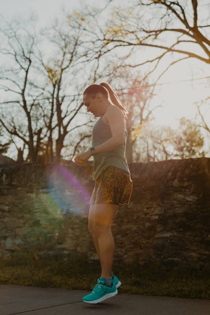 Wearing my running tank top, running skirt, and running shoes, I am doing some pre-run movements to loosen my muscles. I am jumping in the air slightly with my shoes a few inches off the ground. There is a stone wall behind me and the sun is shining bright.