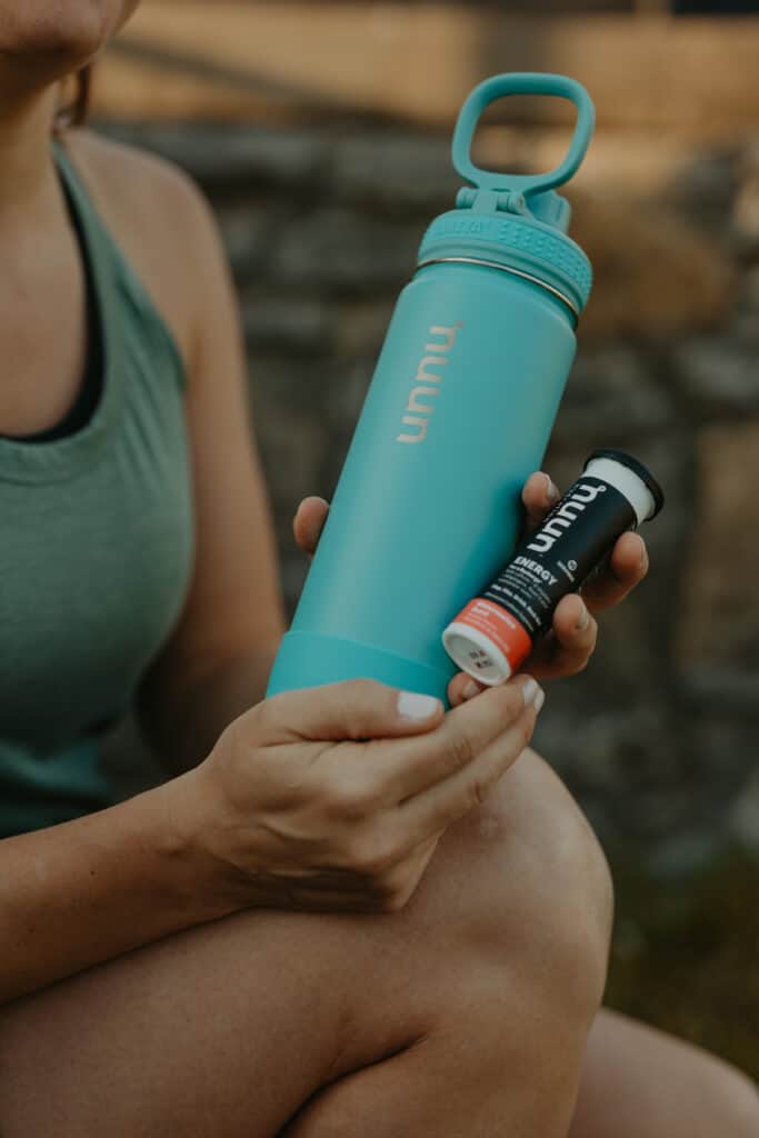 My hands are holding a blue Nuun Hydration branded water bottle and a bottle of Nuun Energy, Watermelon Burst flavor.