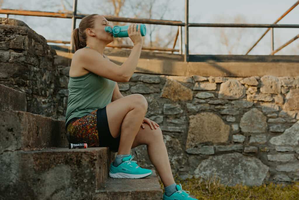 I'm sitting on concrete steps in a park next to a stone wall drinking from a blue Nuun hydration bottle. I'm wearing my running clothes and beside me on the step is a tube of Watermelon Burst Nuun Energy.