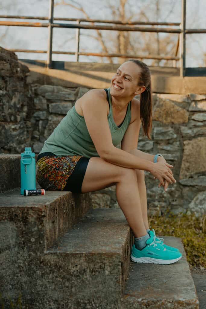 I'm sitting on steps in a park in front of a stone wall. I am looking over my shoulder with a big smile on my face. I'm wearing my running clothes and beside me is a Nuun branded water bottle and a tube of Nuun Energy.