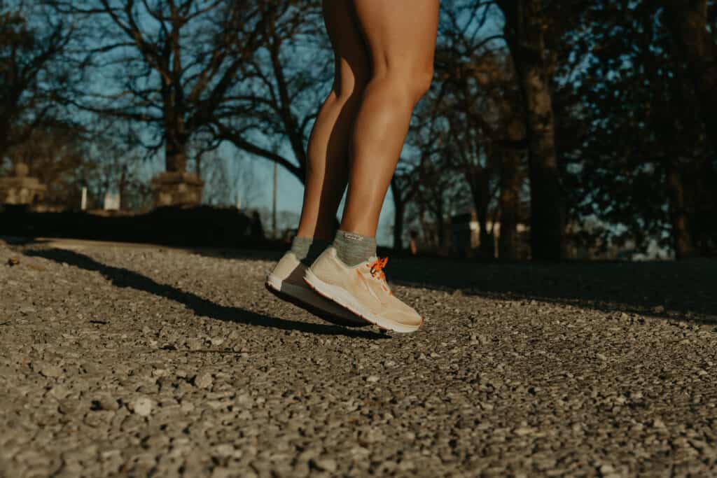 From my thighs down, I am jumping about an inch off the ground. I'm wearing my running socks and running shoes. I am jumping on a gravel path and there are trees all around in the background.