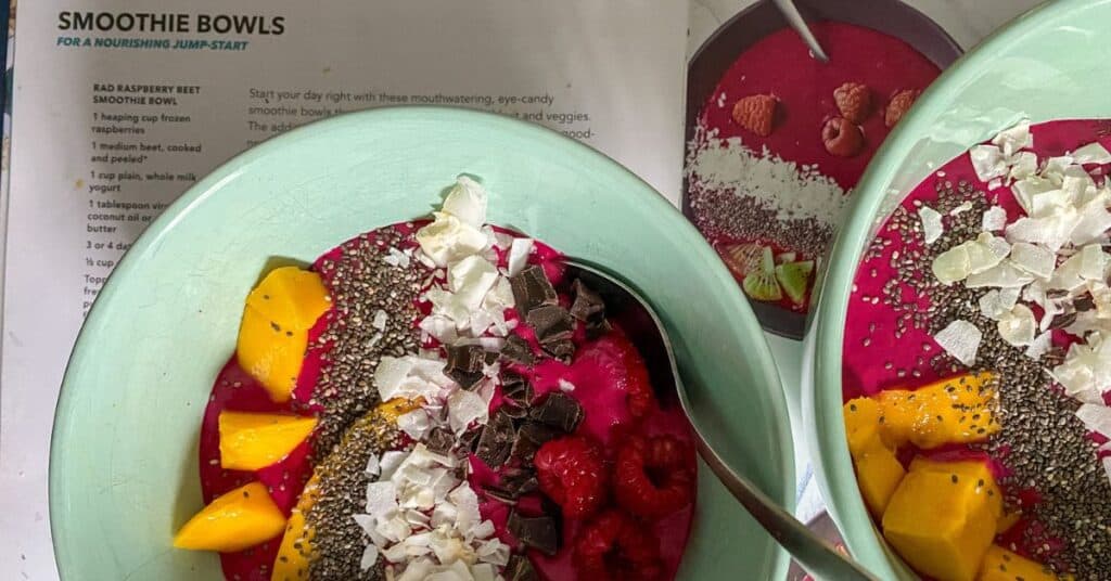 Two bowls are full of red smoothie with fruit, chocolate, coconut flakes, and chia seeds. The bowls are resting on a recipe book with the recipe for the smoothie bowls partially visible.