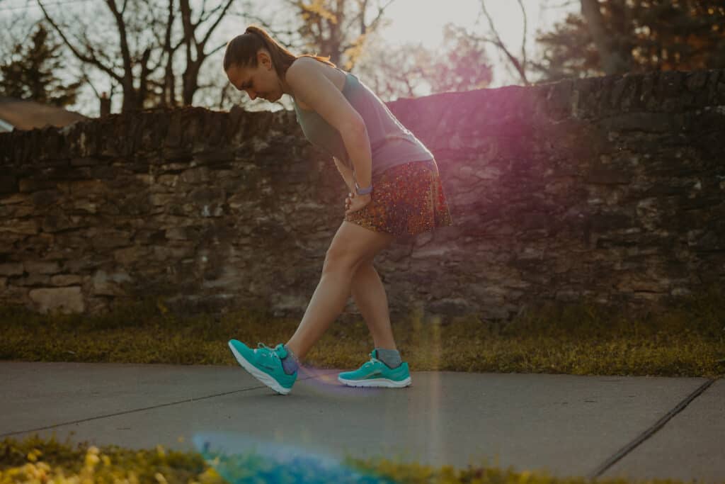 I'm wearing my running clothes in a park with a stonewall behind me. I'm standing on a sidewalk and stretching my hamstring on my left leg by resting my foot on the heel, lifting my toes upward, and leaning my torso over my leg. My hands are both resting on my left leg.