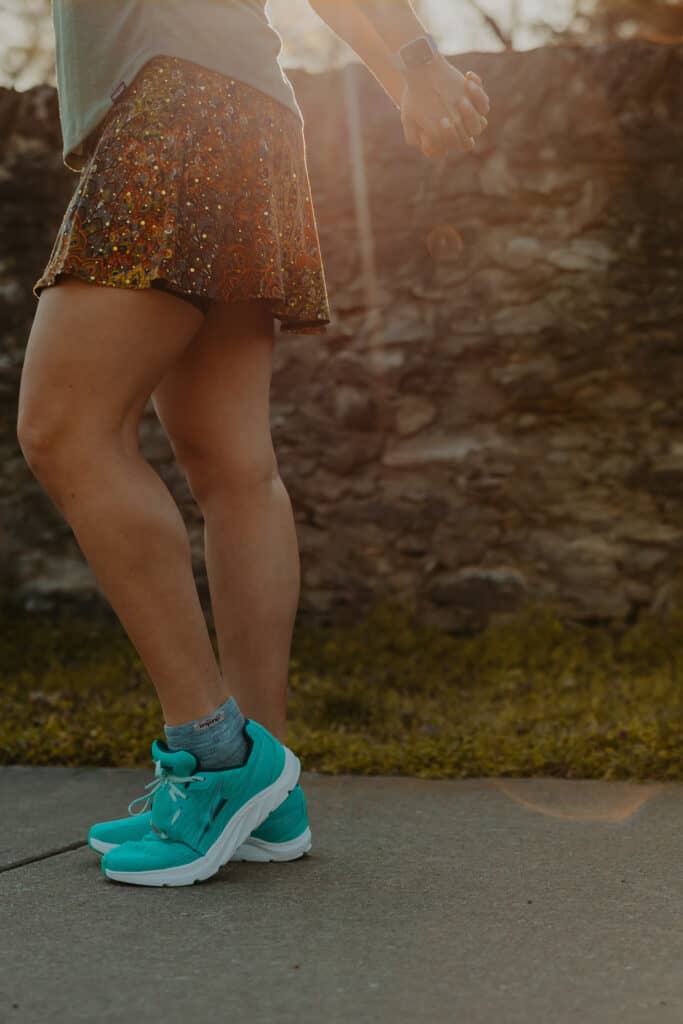 A view from my waist down, I am wearing my running clothes including running skirt and my altra running shoes. My hands are clasped behind my back and I'm stretching my arms back.