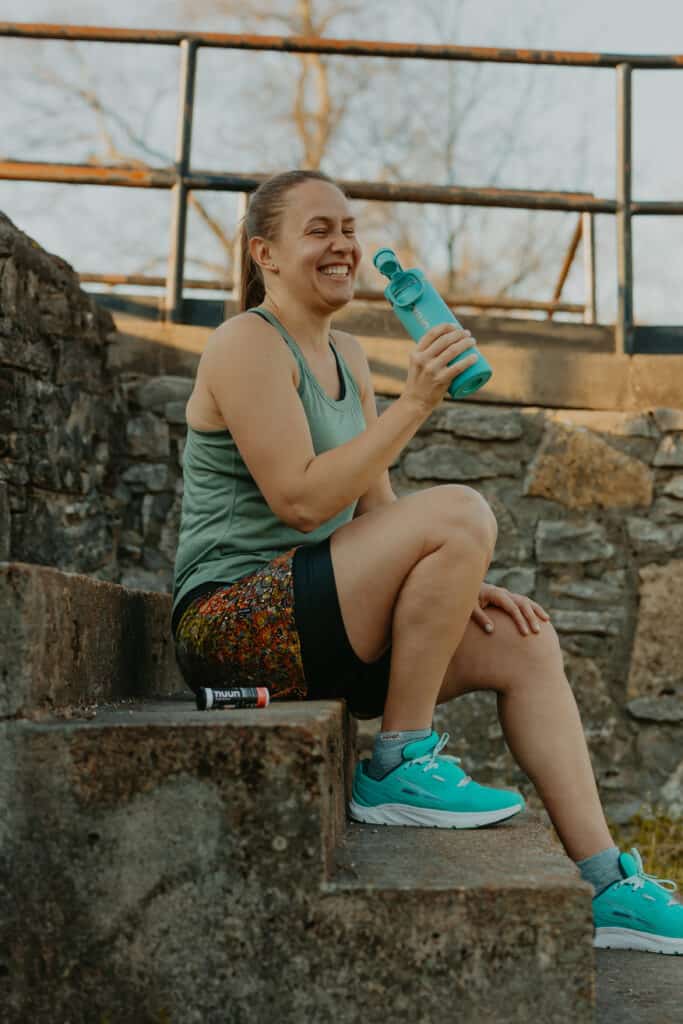 I'm sitting on concrete steps in a park in front of a stone wall. I am about to take a sip from my Nuun water bottle and a tube of Nuun energy is sitting beside me on the step. Nuun hydration is great for athletes and those looking to find success when walking a marathon.