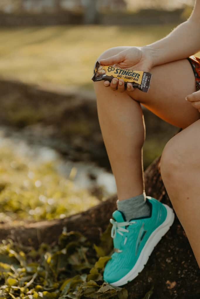 I'm holding a Honey Stinger Oat + Honey bar in my right hand that is resting on my right leg. I'm wearing my running shoes and preparing to run in the park.