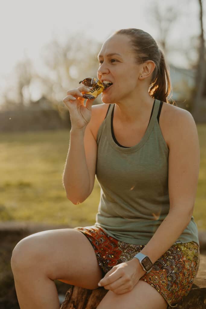 I'm sitting in a park on a tree stump eating a Honey Stinger Oat + Honey bar with the wrapper peeled back a bit. It's a sunny day and I am smiling.