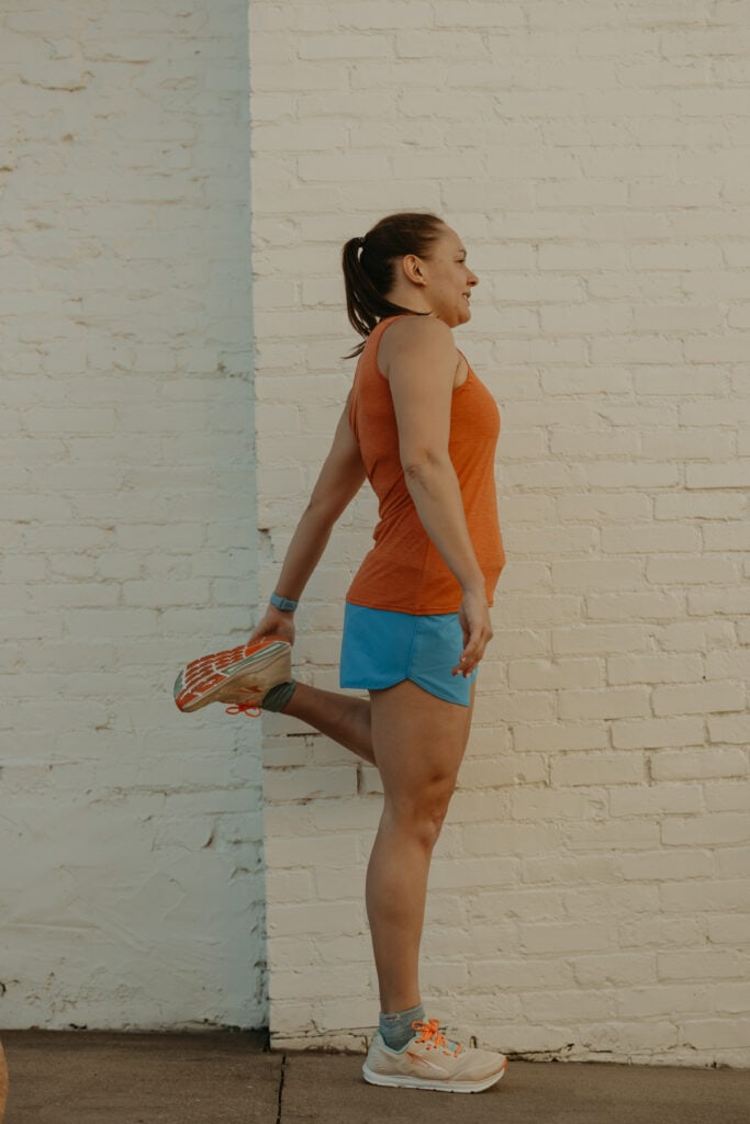 I'm standing beside a white brick wall, stretching my left leg by pulling my foot toward my butt. I'm wearing my running clothes preparing to run.