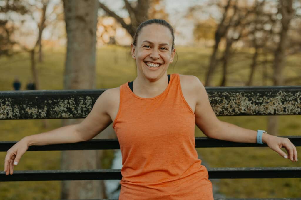 I'm standing in a park wearing my running clothes and leaning up against a railing on a bridge. I have a big smile on my face, and there are many trees in the background.