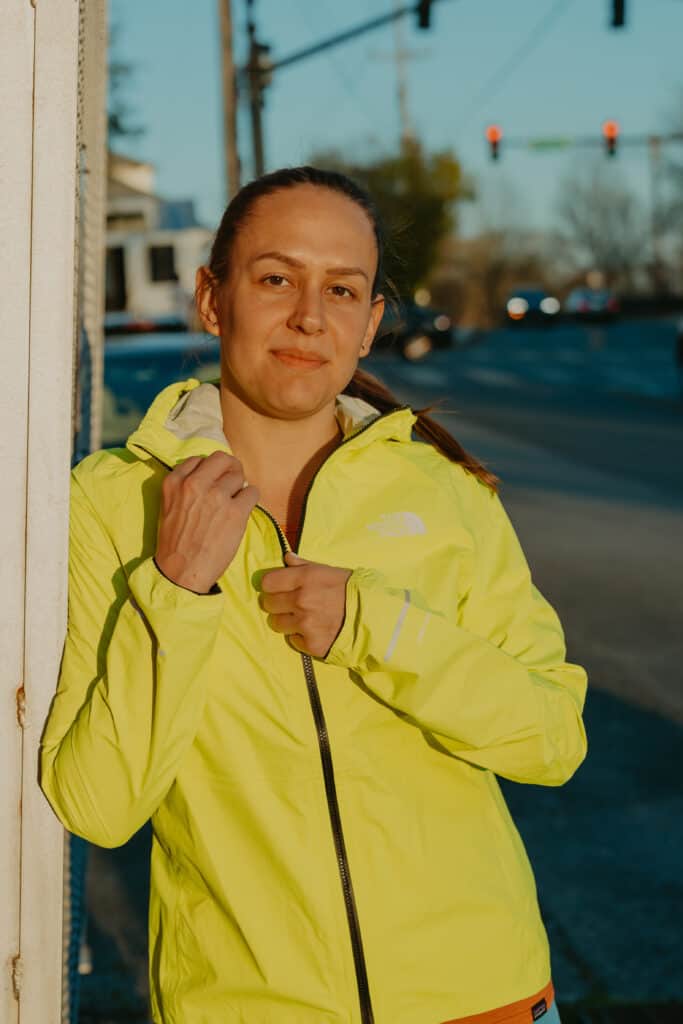 I'm standing leaned up against a building wearing my running rain jacket on a sun evening. I have my hands reaching up for the jacket zipper and collar of the jacket. There is a road behind me out of focus along with some cars and a red traffic light.