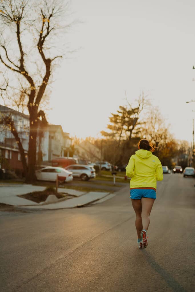 I'm running down a neighborhood street lined with houses and trees as the sun sets in the distance. I am running away from the camera, dressed in my running clothes including a running rain jacket and running shorts.